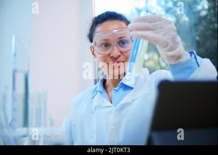 Nahaufnahme einer schönen afroamerikanischen Frau in einer Schutzbrille, die Reagenzgläser mit chemischen Substanzen untersucht, im Biolab Stockfoto