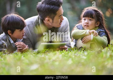 asiatischer Vater, der vorne auf Gras liegt und zwei Kindern glücklich und lächelnd Geschichten erzählt Stockfoto