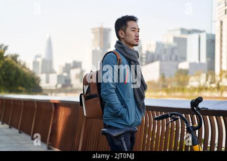 Junger asiatischer Erwachsener mit Rucksack, der an seinem Fahrrad steht und die Skyline der Stadt im Park am Flussufer betrachtet Stockfoto