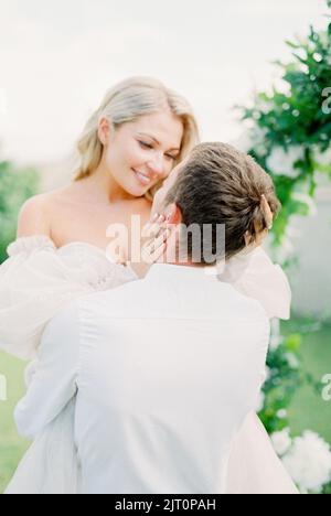 Die Braut berührt das Gesicht des Bräutigams, der sie in seinen Armen hält. Hochformat Stockfoto