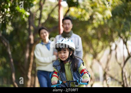 Kleine asiatische Mädchen Reiten Fahrrad im Stadtpark mit Eltern im Hintergrund Stockfoto