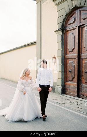 Die Braut mit dem Bräutigam an der Hand geht an der Holztür des Gebäudes vorbei Stockfoto