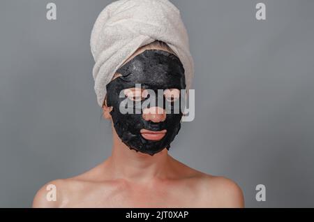 Porträt einer Frau in weißem Handtuch auf dem Kopf und schwarzer Stoffmaske auf ihrem Gesicht. Feuchtigkeitsspendende Haut. Grauer Hintergrund. Stockfoto