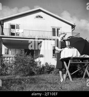 Mutter mit Kind im Jahr 1940s. Eine junge Mutter im Garten mit ihrem Haus im Hintergrund schaut auf ihr Neugeborenes, das in einer Kratsche liegt und einen Regenschirm zusammengelegt hat, um vor dem Sonnenlicht zu schützen. Schweden 1947 Kristoffersson Ref. V94-3 Stockfoto