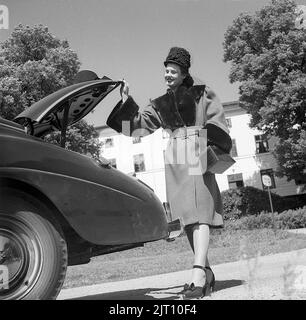 Damenmode im Jahr 1940s. Eine junge Frau in einem typischen Outfit von 1940s. Ein warmer Mantel, passende Schuhe, Handtasche und Hut. Sie posiert das Outfit neben einem Auto und einem offenen Kofferraum. Schweden 1946. Kristoffersson Ref. U129-3 Stockfoto