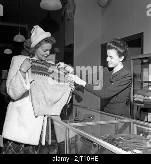 Einkaufen in der 1940s. Eine junge Frau in einem Geschäft, in dem eine Verkaufsfrau einige der Geschäfte mit warmen Winterkleidung präsentiert, ist ein Strickpullover von besonderem Interesse und hält ihn vor sich, wobei das schöne Strickmuster sichtbar ist. Schweden 1947. Kristoffersson Ref. Y71-3 Stockfoto