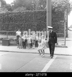 1950s Polizist. Er weist eine Gruppe von Kindern an, wie man eine geschäftige Straße auf sichere Weise überquert. Die Kinder befolgen seine Anweisungen sorgfältig, schauen beide Richtungen, bevor sie die Straße überqueren. Sie wechseln sich ab, um mit dem Polizisten zu gehen. Das Jahr ist 1958. Stockfoto