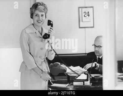 Büromädchen in den 1960s. Eine Frau steht in einem Büro mit einem Stift und einem Notizbuch, das ein Telefon hält. Man sieht einen Mann an einem Schreibtisch sitzen. Sie ist Sekretärin. Schweden 1961 Stockfoto