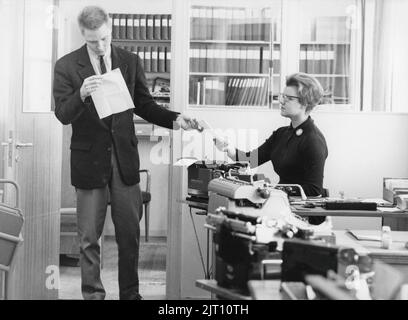 Büromädchen in den 1960s. Eine Frau sitzt neben ihrer Schreibmaschine und ein Mann steht im Raum, der gleichzeitig ein Dokument von ihr liest und aushändigt. Ein gutes Beispiel dafür, wie die Beziehungen zwischen Mitarbeitern und Managern damals waren, da er ihr weder Aufmerksamkeit noch Dankbarkeit für ihre Arbeit schenkt, sichtbar ein Mann, der sich selbst als auf einer höheren Ebene im Unternehmen sieht als sie. Schweden 1960. Stockfoto