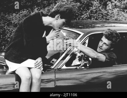 In der 1960s. Eine Frau sitzt auf der Motorhaube eines Autos, und ein Mann greift mit einem Zigarettenanzünder, um sie anzuzünden. Das Jahr ist 1964. Schweden Stockfoto