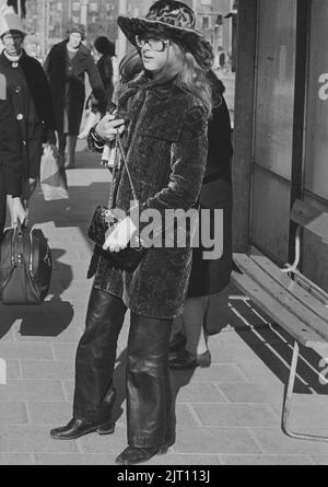 In der 1970s. Eine junge Frau auf der Straße in Lederhosen, halblangem Samtmantel und passendem Hut. Die Gläser sind typisch 1970s. Das Jahr ist 1970. Stockfoto