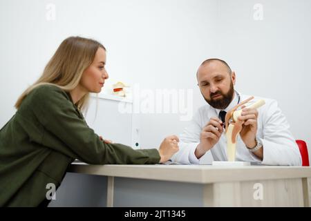 Hübsche junge Patientin bei Termin in einem Orthopäden oder Traumatologen. Physiotherapeut Arzt mit Kniegelenkmodell Stockfoto