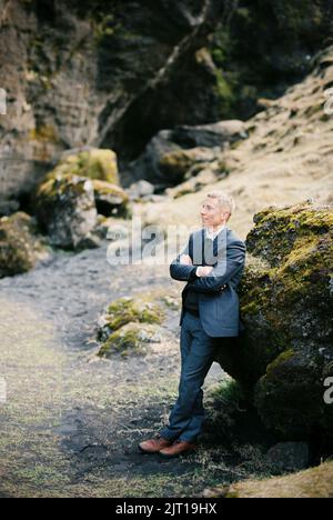 Der Mann in einem Anzug mit gekreuzten Armen auf der Brust steht auf einem riesigen Felsbrocken gelehnt Stockfoto