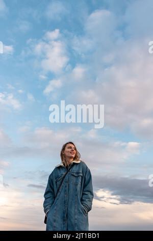 Eine blonde Frau, die in einem blauen Mantel mit dem blauen Himmel im Hintergrund wegschaut Stockfoto