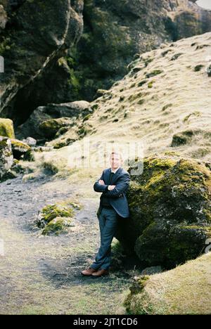 Der Mann in einem Anzug steht in der Nähe eines riesigen, mit Moos bedeckten Felsens. Island Stockfoto