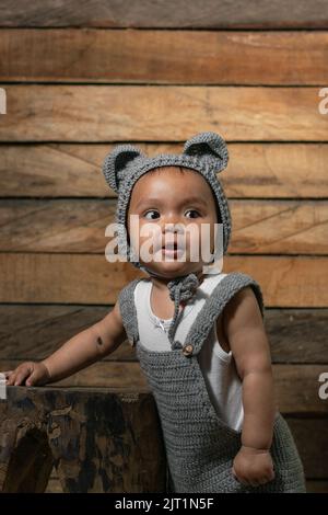 Schöne Baby Latina mit brauner Haut, auf einem Holzstuhl und einem orangen Hintergrund stehend, lernen, ihre ersten Schritte zu machen, Overalls und ein c tragen Stockfoto