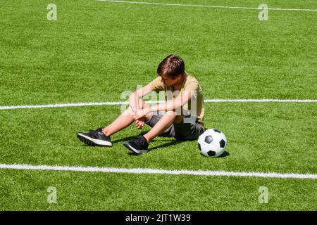 Trauriger, allein jugendlicher Junge, der im leeren Schulsportstadion im Freien sitzt. Emotionen, Niederlage, verlorenes Spiel, Schwierigkeiten, Probleme von Jugendlichen Stockfoto