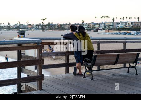 Ein Paar auf einem hölzernen Pier, das den Strand bei Sonnenuntergang beobachtet Stockfoto