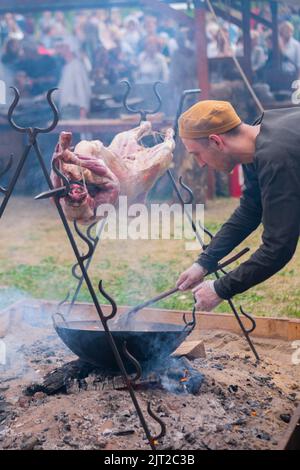 Mann, der über offenem Feuer gebratenes Schweinefleisch auf Spucken und Gemüse im Topf kocht Stockfoto