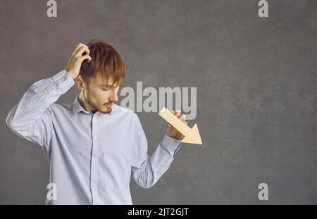 Ein junger Mann mit einem rätselhaften Ausdruck sieht auf einen hölzernen Pfeil in seinen Händen, der nach unten zeigt. Stockfoto