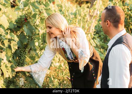 Junge blonde Frau in serbischer traditioneller Kleidung, die mit ihrem Mann grüne Trauben anschaut Stockfoto