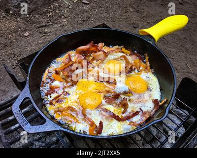 Ein köstliches Frühstück während des Campings mit Eiern und Speck, der in einer eisernen Pfanne über Holzkohle brutzelt Stockfoto