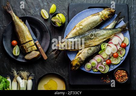 Mediterrane Küche, geräucherter Heringsfisch serviert mit grünen Zwiebeln, Zitrone, Kirschtomaten, Gewürzen, Brot und Tahini-Sauce auf dunklem Hintergrund. Stockfoto