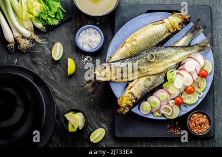 Mediterrane Küche, geräucherter Heringsfisch serviert mit grünen Zwiebeln, Zitrone, Kirschtomaten, Gewürzen, Brot und Tahini-Sauce auf dunklem Hintergrund. Stockfoto