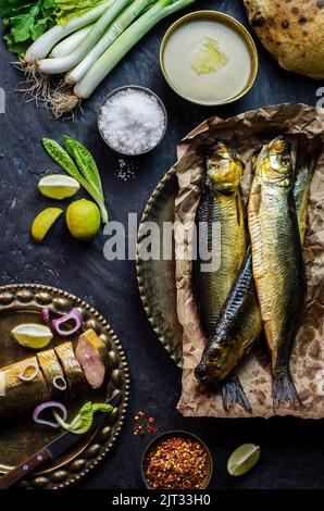 Mediterrane Küche, geräucherter Heringsfisch serviert mit grünen Zwiebeln, Zitrone, Kirschtomaten, Gewürzen, Brot und Tahini-Sauce auf dunklem Hintergrund. Stockfoto