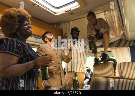 Interracial Gruppe von engen Freunden tanzen zur Musik und singen in ihrem komfortablen geräumigen Wohnmobil, und Bier trinken. Wir feiern im Sommer einen Geburtstag auf einer Campingbus-Reise. Hochwertige Fotos Stockfoto