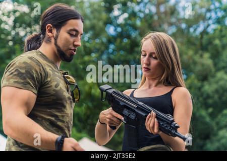 Männlicher kaukasischer muskulöser Schießlehrer im moro-T-Shirt, der seiner Kundin die Gewehre zeigt. Konzept der Pistolenreihe. Unkonventionelles Hobby. Hochwertige Fotos Stockfoto