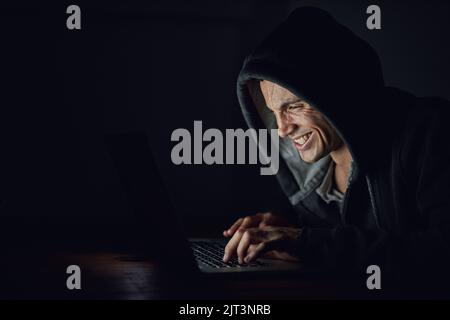 Dunkle Taten in einer dunklen Nacht. Ein kapuzter Computerhacker, der im Dunkeln einen Laptop benutzt. Stockfoto
