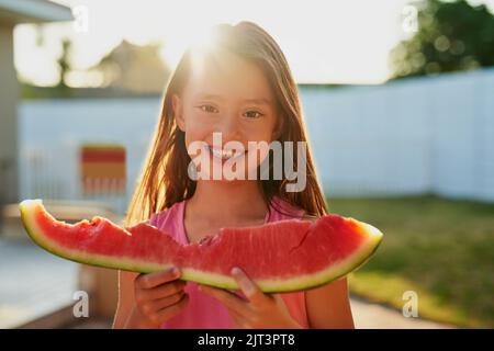 Der erste Geschmack des Sommers. Porträt eines glücklichen jungen Mädchens, das im Hinterhof ein Stück Wassermelone isst. Stockfoto