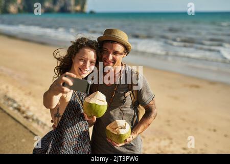 Strandtage mit meinem Beach Babe. Ein glückliches junges Paar, das Selfies macht, während es Cocktails am Strand genießt. Stockfoto