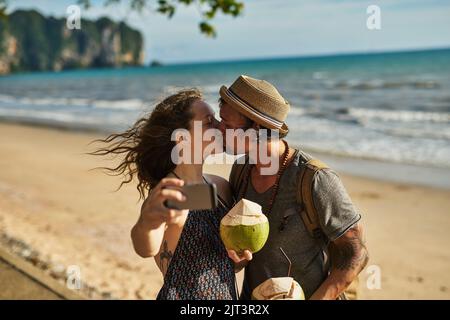 Küsse sind flüchtig, aber Selfies sind ewig. Ein glückliches junges Paar, das Selfies macht, während es Cocktails am Strand genießt. Stockfoto