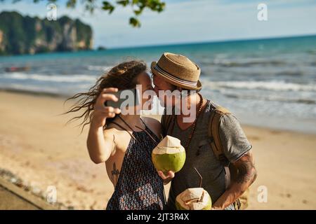 Sommerzeit ist Selfie-Zeit. Ein glückliches junges Paar, das Selfies macht, während es Cocktails am Strand genießt. Stockfoto