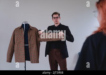 Eleganter Modedesigner. Kaukasischer blonder Mann, der in einem schwarzen Hemd, einer braunen Hose und einer Brille als Schneider arbeitet. Hochwertige Fotos Stockfoto