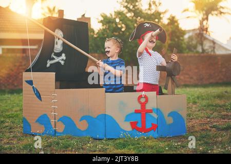 Auf der Jagd nach vergrabenen Schätzen. Ein süßer kleiner Junge und sein Bruder spielen Piraten draußen auf einem Boot aus Pappkartons. Stockfoto