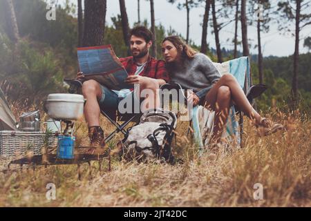 Junges Paar, das sich auf einem Stadtplan ansieht, während es auf seinem Campingplatz sitzt. Abenteuerliches junges Paar, das im Wald zeltet. Stockfoto