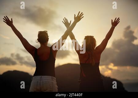 Die Hände zum Sonnenuntergang erheben. Rückansicht von zwei jungen Freunden mit erhobenen Armen, die vor einem Sonnenuntergang gedreht wurden. Stockfoto