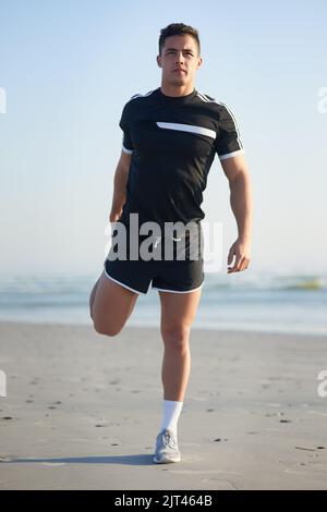 Ich fange immer mit einem Warm-up an. Porträt eines hübschen jungen Mannes, der sich vor einem Workout am Strand aufwärmt. Stockfoto