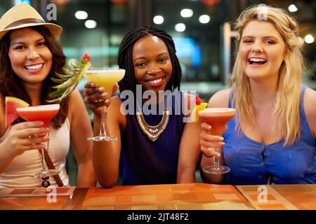 Getränke an der Bar. Beschnittenes Porträt von drei jungen Freundinnen, die an einer Bar einen Drink genießen. Stockfoto