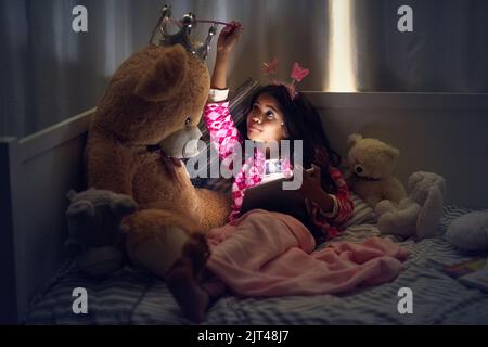 Kinder genießen es, jeden Moment magisch zu machen. Ein kleines Mädchen benutzt ein digitales Tablet, während es mit ihrem Teddy nachts im Bett liegt. Stockfoto