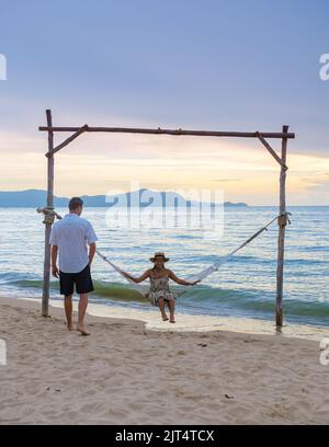 Männer und Frauen beobachten den Sonnenuntergang von einer Hängematte am Strand in Pattaya Thailand Ban Amphur Strand. Paar zu Fuß auf einem tropischen Strand mit Palmen und Hängematte während des Sonnenuntergangs Stockfoto