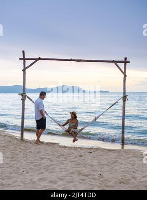 Männer und Frauen beobachten den Sonnenuntergang von einer Hängematte am Strand in Pattaya Thailand Ban Amphur Strand. Paar zu Fuß auf einem tropischen Strand mit Palmen und Hängematte während des Sonnenuntergangs Stockfoto