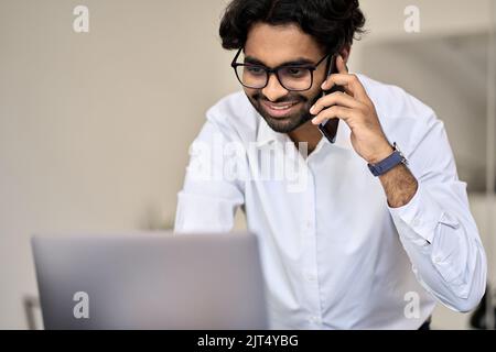 Lächelnder indischer Geschäftsmann, der am Telefon telefoniert und im Büro einen Laptop anschaut. Stockfoto
