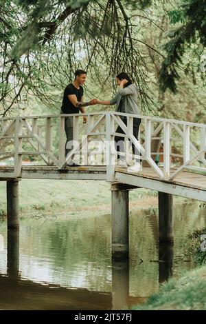 Schönes Paar, das sich die Hände schüttelt, während es auf der weißen Brücke im Park steht Stockfoto