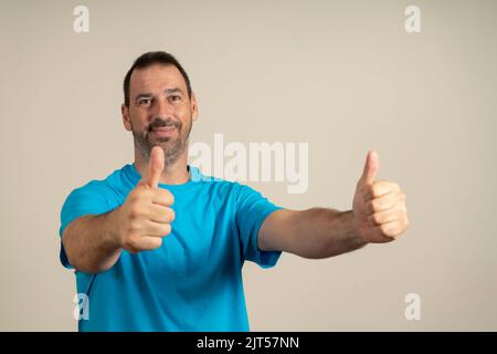 Attraktiver, fröhlicher mann mit lateinischem Bart, der ein blaues T-Shirt trägt, das isoliert auf beigem Hintergrund steht und die Daumen nach oben zeigt Stockfoto