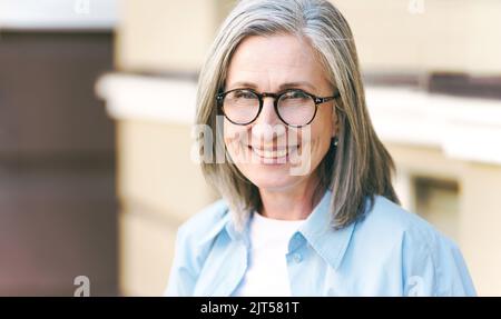 Charmante graue Haare reife Frau trägt eine Augenbrille im Freien stehen genießen Urlaub rund um die Welt im Ruhestand reisen. Reife Frau mit perfekter Haut trägt blaues Hemd. Stockfoto