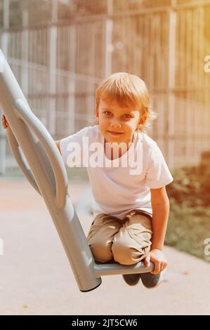 Kind auf Stadtspielplatz Schaukeln auf Schaukel Karussell. Nette kleine glücklich lächelnd offen fünf Jahre alten Jungen mit langen blonden Haaren in einem weißen T-Shirt. gen Stockfoto